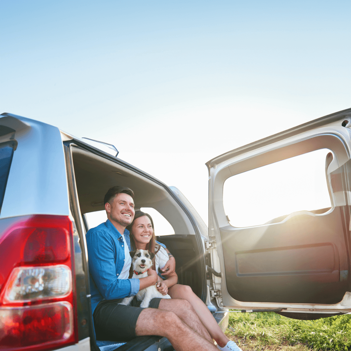 happy couple traveling with dog sitting in back of their car