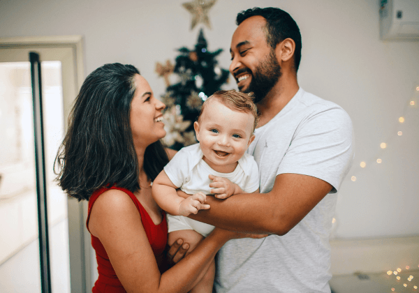 a man and woman smiling holding their baby in front of their christmas tree