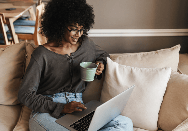 woman drinking coffee smiling at her laptop on her living room couch